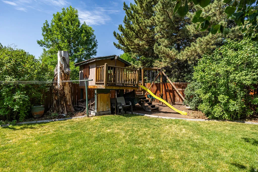 Expansive backyard featuring a lush lawn, mature trees, wood fence, and a multi-level playset with a yellow slide
