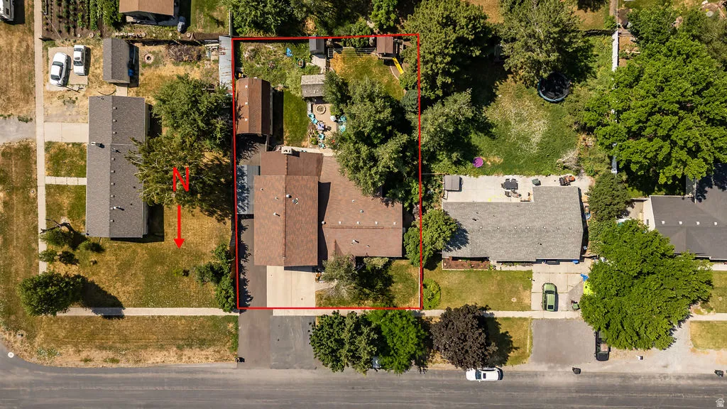Aerial view showcasing a residential property with a brown-roofed home, a paved driveway, and a spacious backyard featuring mature trees and a patio area