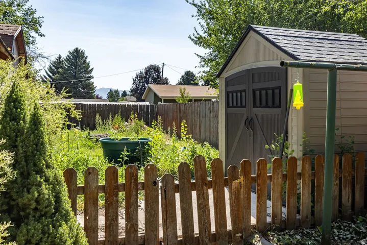 Garden space featuring a plastic shed with double doors, wood picket fencing, and mature evergreen trees
