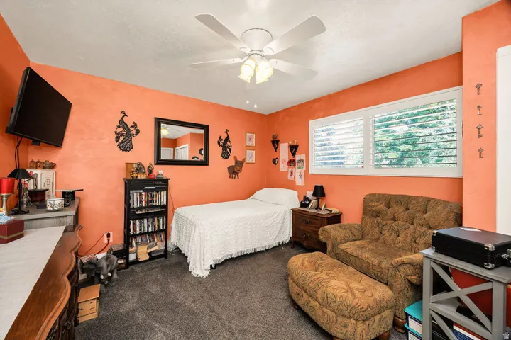 Interior room featuring textured orange walls, gray carpet, a white ceiling fan with integrated lighting, and a large window with white plantation shutters