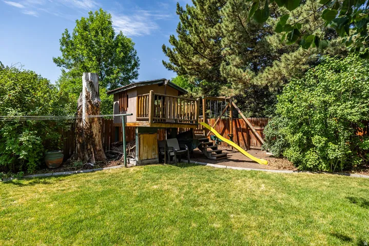Expansive backyard featuring a lush lawn, mature trees, wood fence, and a multi-level playset with a yellow slide