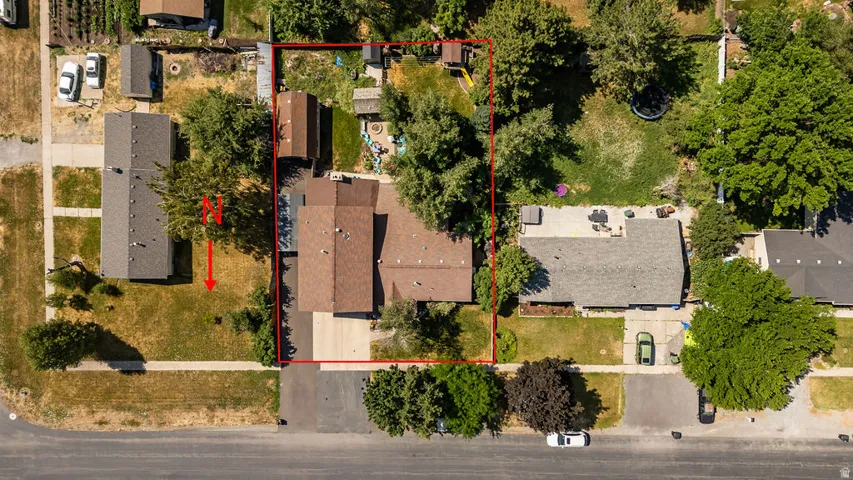 Aerial view showcasing a residential property with a brown-roofed home, a paved driveway, and a spacious backyard featuring mature trees and a patio area