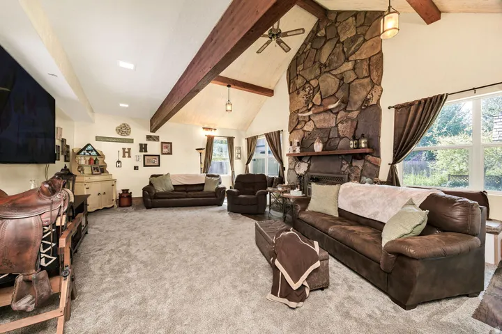 Spacious living area featuring a vaulted ceiling with exposed wood beams, a stone-clad fireplace, and large windows