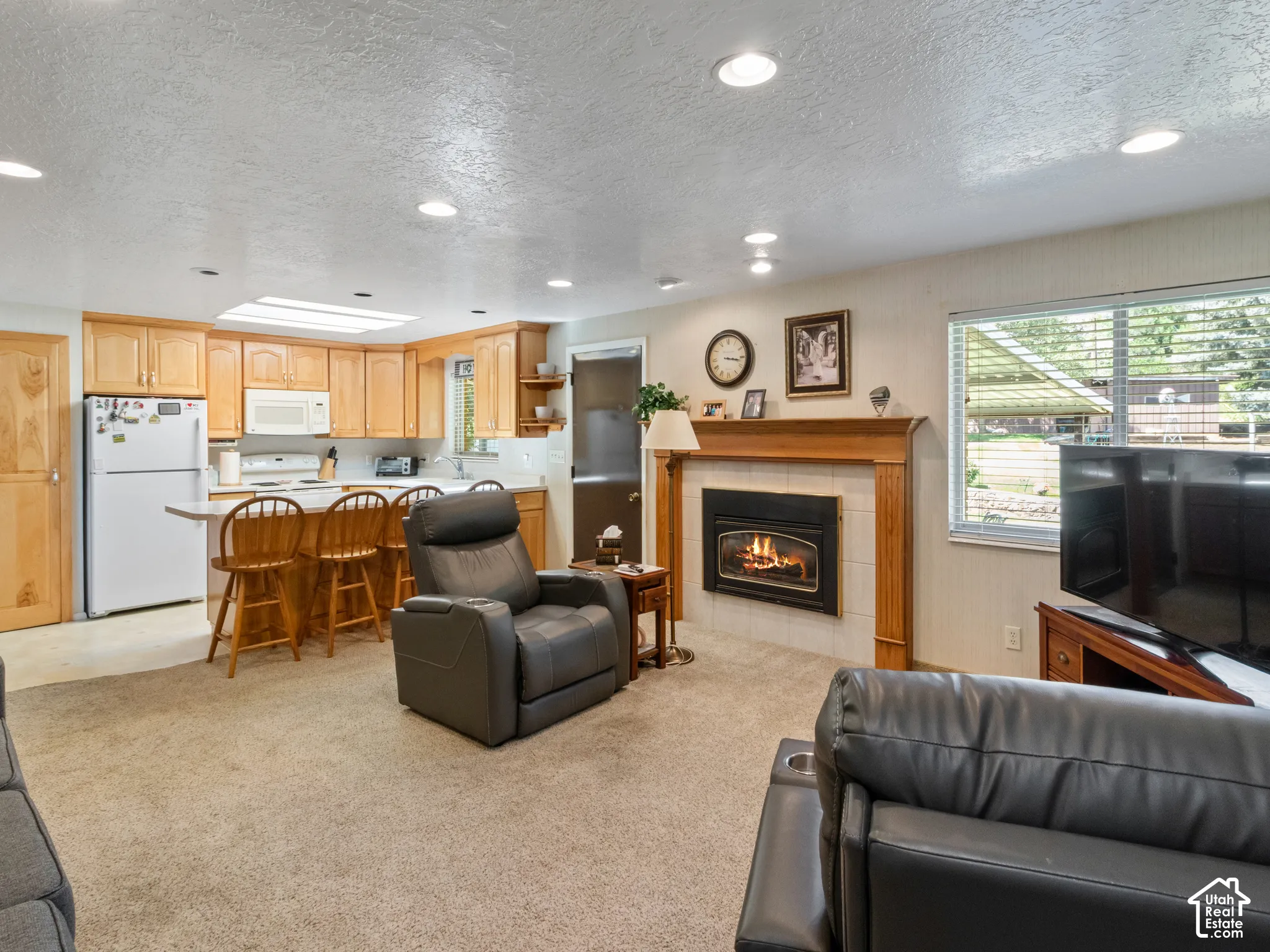 Living area featuring a gas fireplace with a wood mantle and tile surround, recessed lighting, and a kitchen with wood cabinetry and a breakfast bar