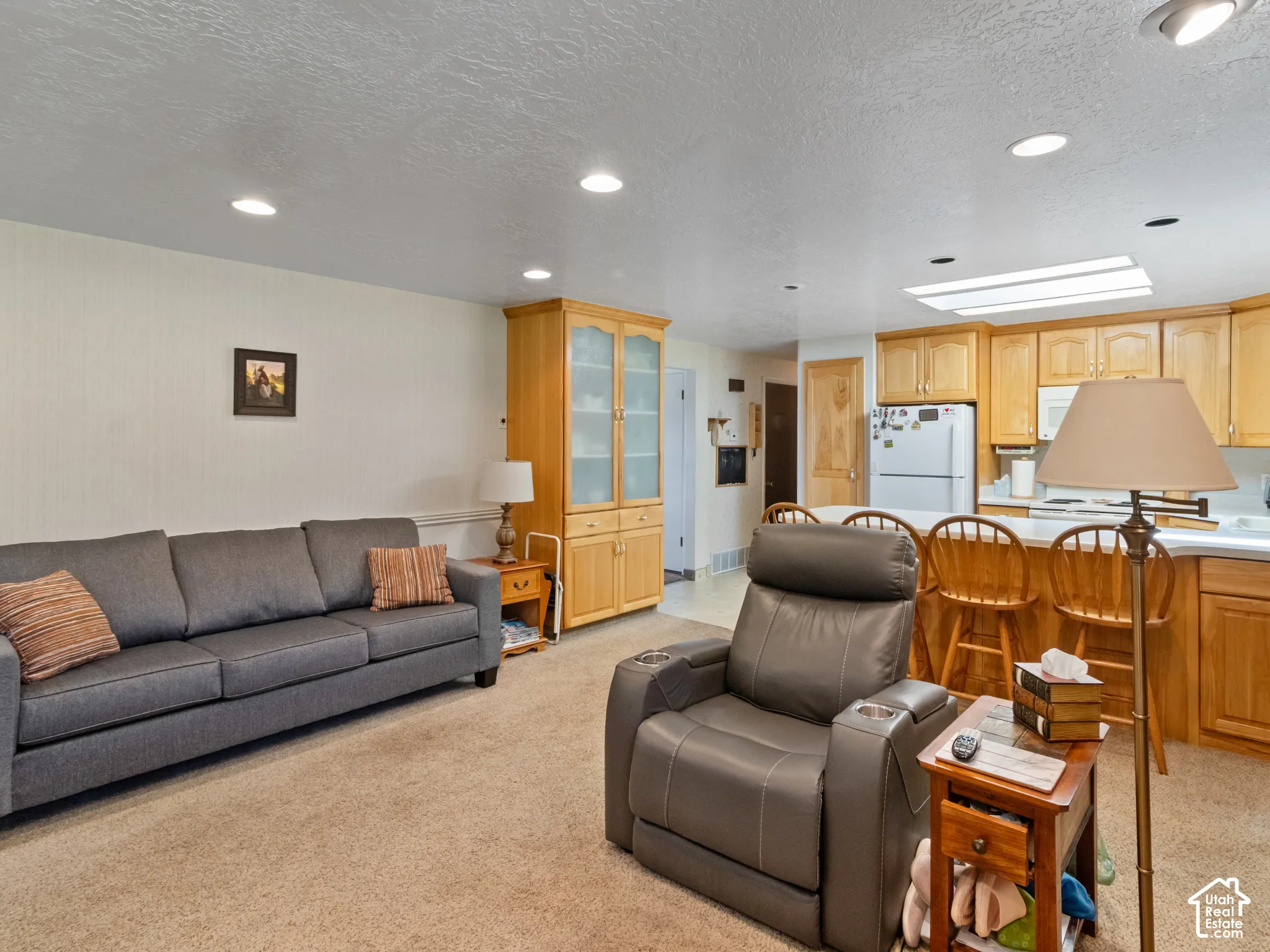 Open-concept living space featuring recessed lighting, plush carpet flooring, a built-in display cabinet with glass doors, a kitchen island with seating, and a full suite of kitchen appliances