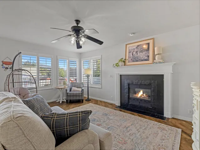 Living area featuring a fireplace with a white mantel and black tile surround, wood-finish flooring, a ceiling fan, and multiple windows with white plantation shutters