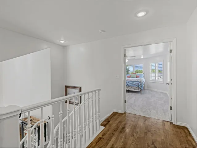 Hardwood-finish flooring leading to a bedroom featuring light gray carpeting, two windows with white plantation shutters, and a ceiling fan