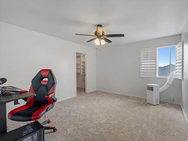 Carpeted interior space featuring a window with white louvered shutters, a ceiling fan with integrated lighting, and an open doorway revealing a closet with built-in shelving