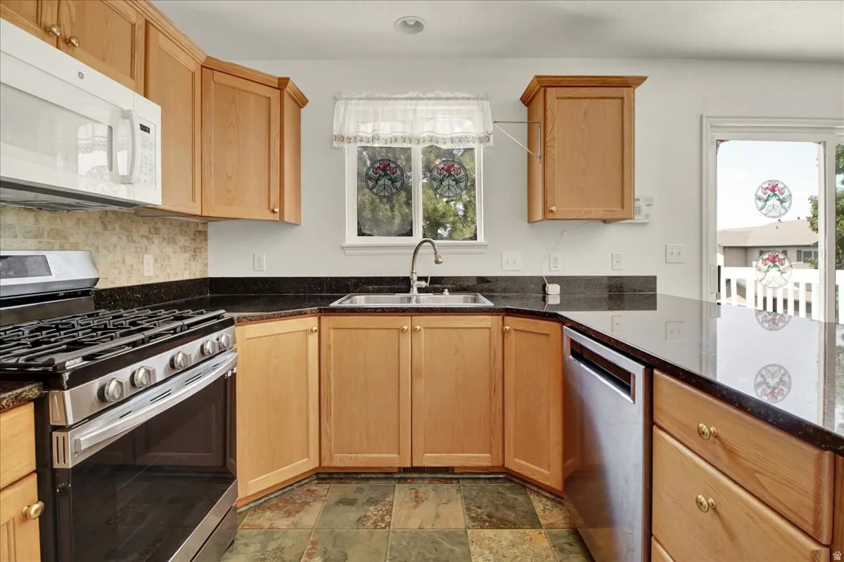 Kitchen featuring light wood cabinetry, dark granite countertops, stainless steel appliances, and a mosaic tile backsplash