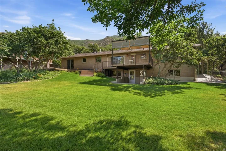 Expansive green lawn with mature trees and a two-story residence featuring a brick facade, upper-level deck with shade covering, and a lower-level patio