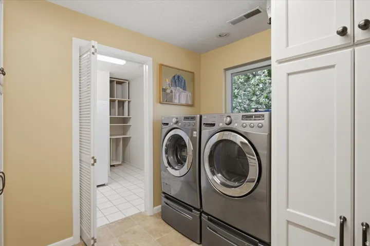 Laundry area featuring neutral wall paint, tiled flooring, white cabinetry with dark hardware, a window offering natural light, and an adjacent storage space with shelving and white tile flooring