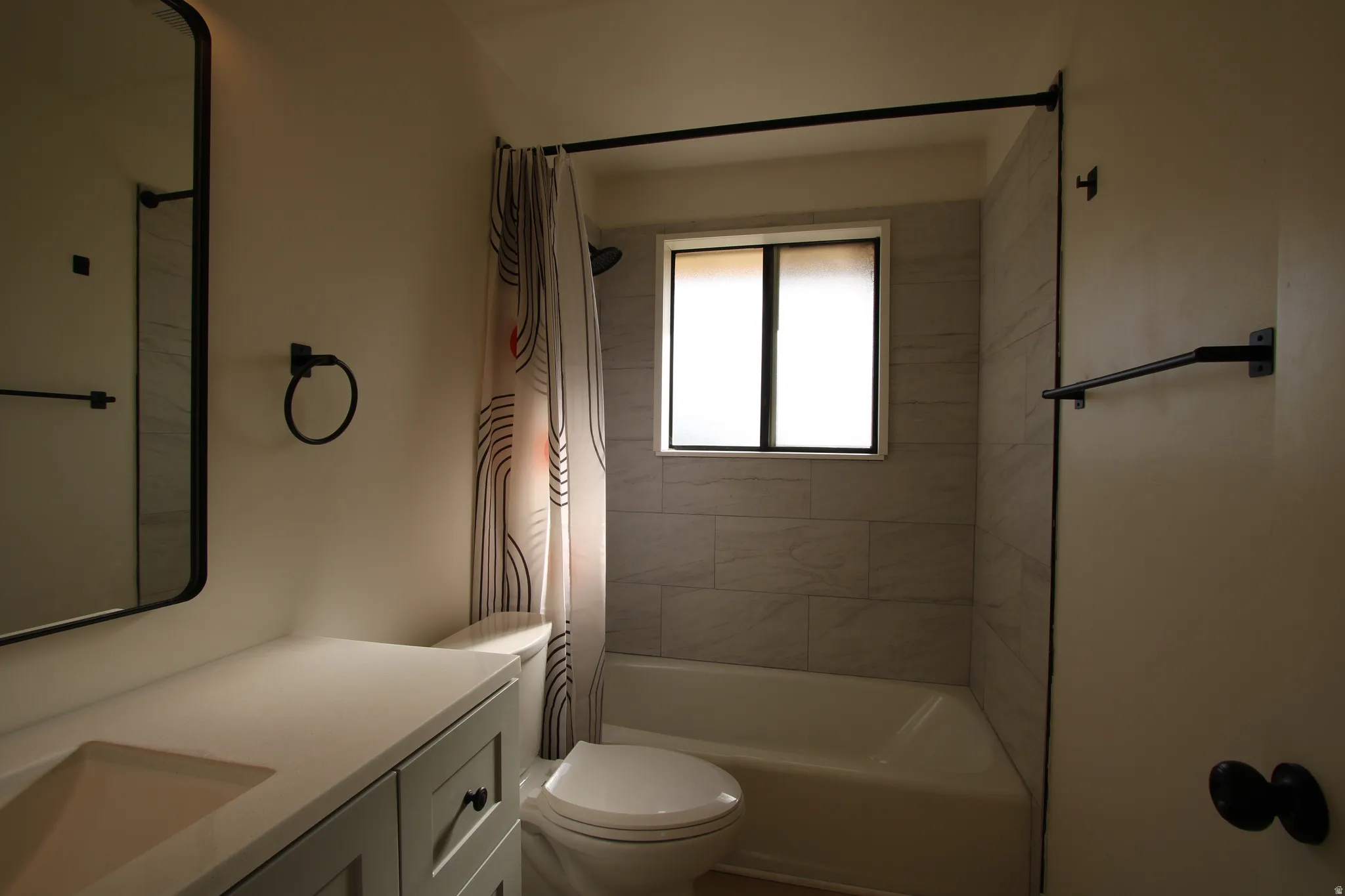 Bathroom featuring a built-in tub with tile surround, a window with privacy blind, a vanity with an integrated sink and light-toned countertop, and matte black hardware