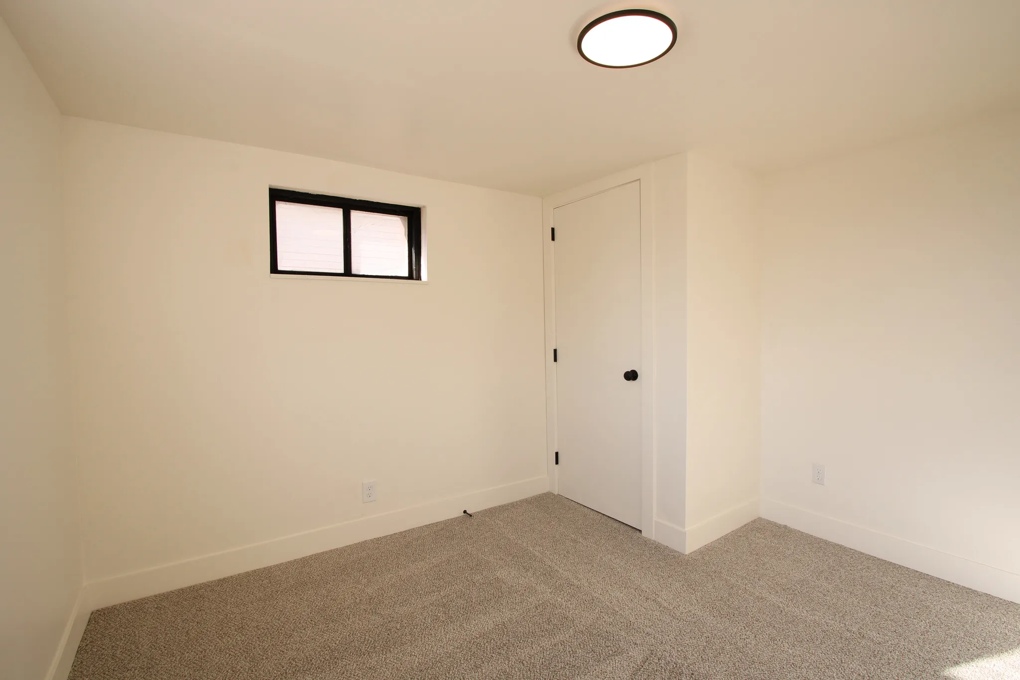 Room featuring neutral-toned walls, gray textured carpeting, a flush-mount ceiling light fixture, a white door with black hardware, and a window with black trim