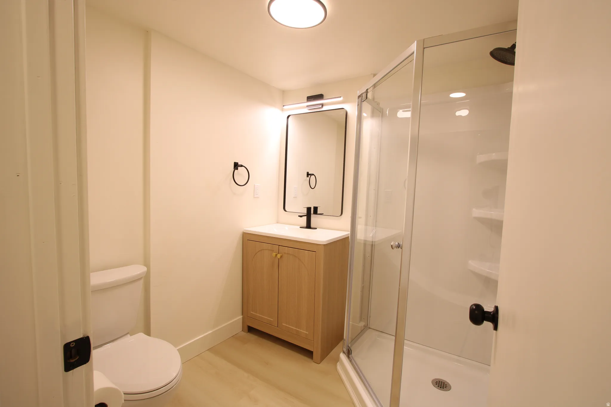 Modern bathroom featuring a glass-enclosed shower with integrated shelving, a light wood-finish vanity with a white countertop, and a contemporary black-framed mirror