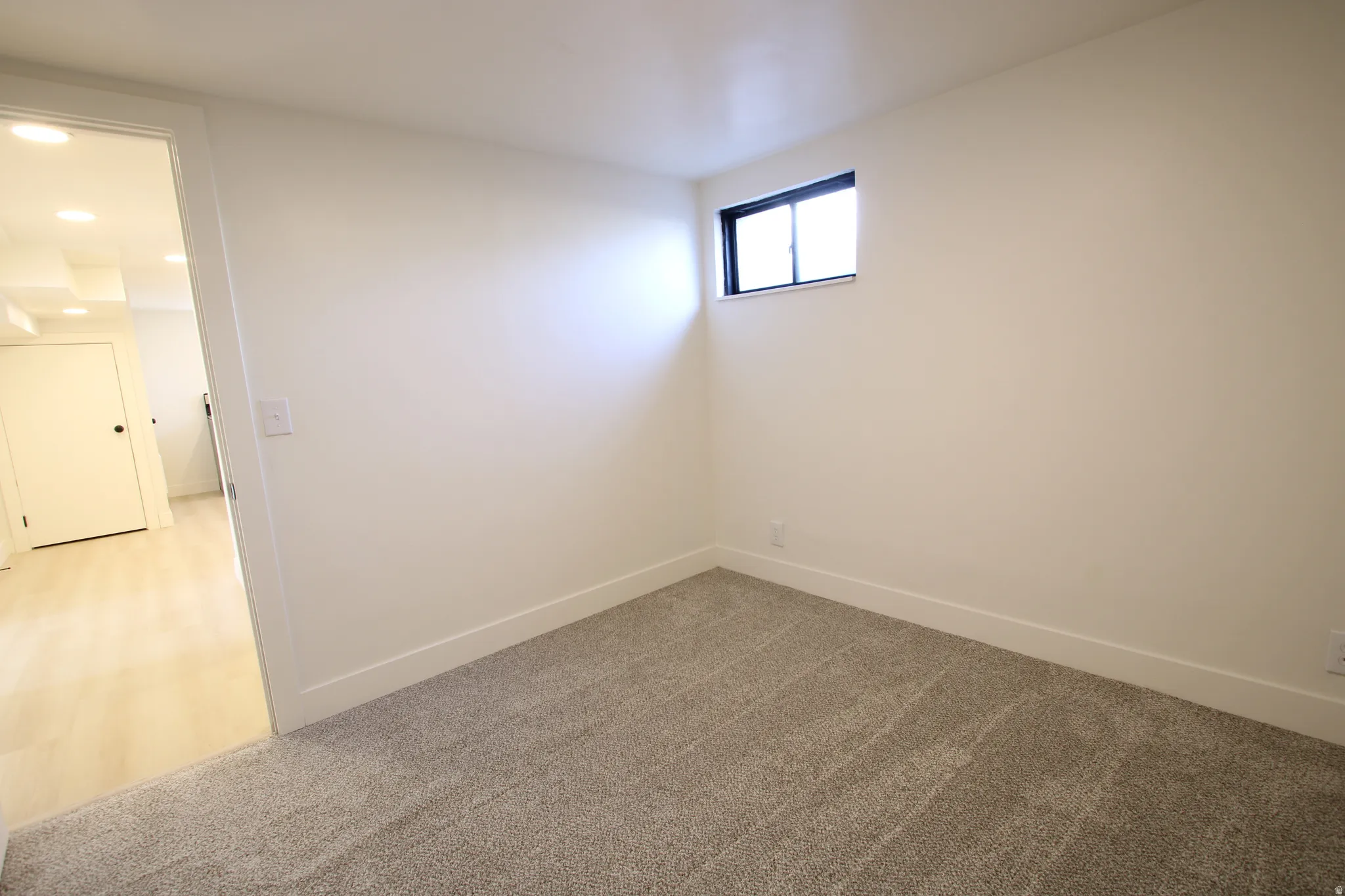 Room featuring neutral carpeting, white painted walls, a small window with a dark frame, and white baseboards