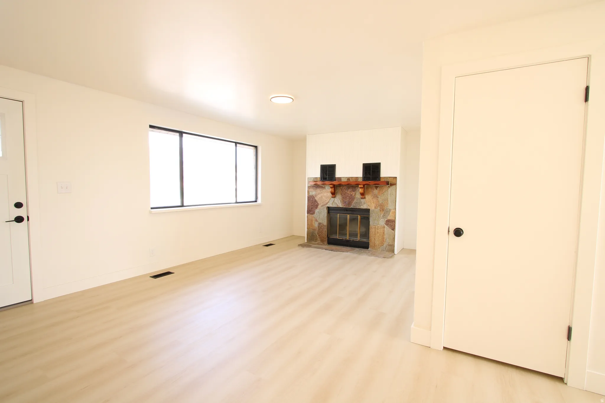 Spacious room featuring light wood-finish flooring, a stone-clad fireplace with a wood mantel, a large window with dark trim, a white entry door with black hardware, and a ceiling-mounted light fixture