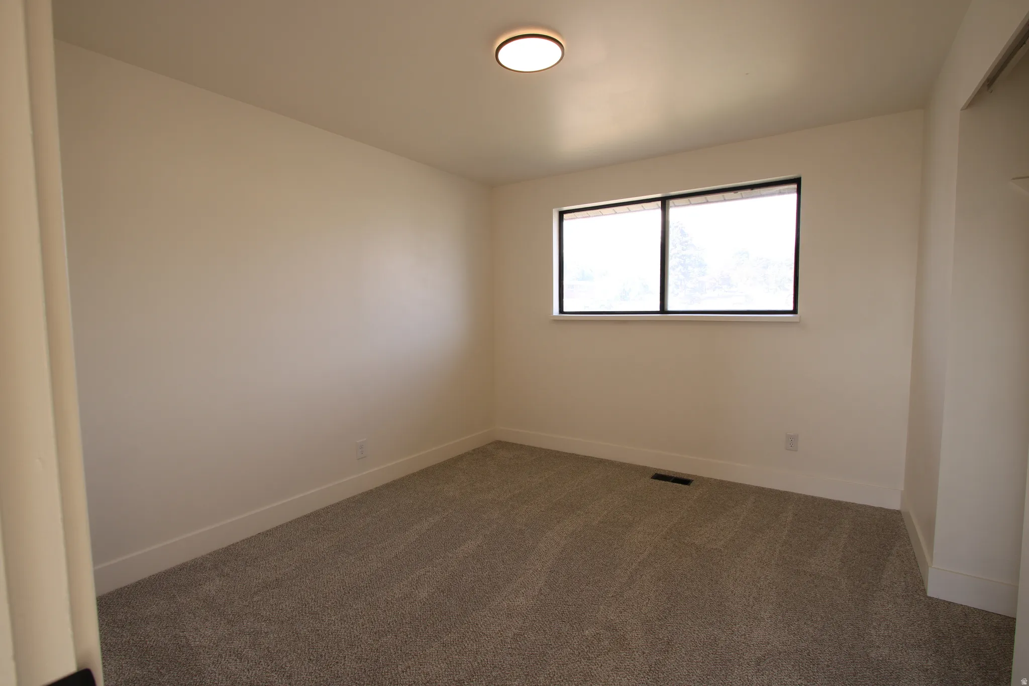 Interior room featuring light-toned walls, grey carpet flooring, a single window with dark framing, and a flush-mount ceiling light fixture