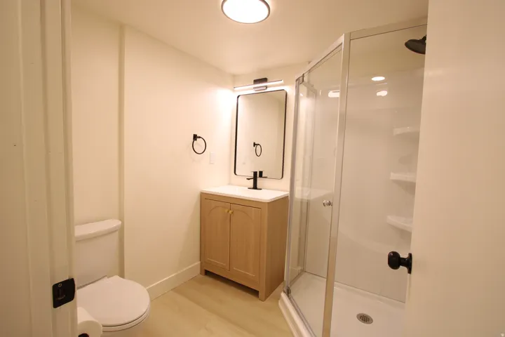 Modern bathroom featuring a glass-enclosed shower with integrated shelving, a light wood-finish vanity with a white countertop, and a contemporary black-framed mirror