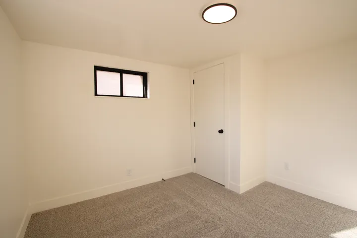 Room featuring neutral-toned walls, gray textured carpeting, a flush-mount ceiling light fixture, a white door with black hardware, and a window with black trim