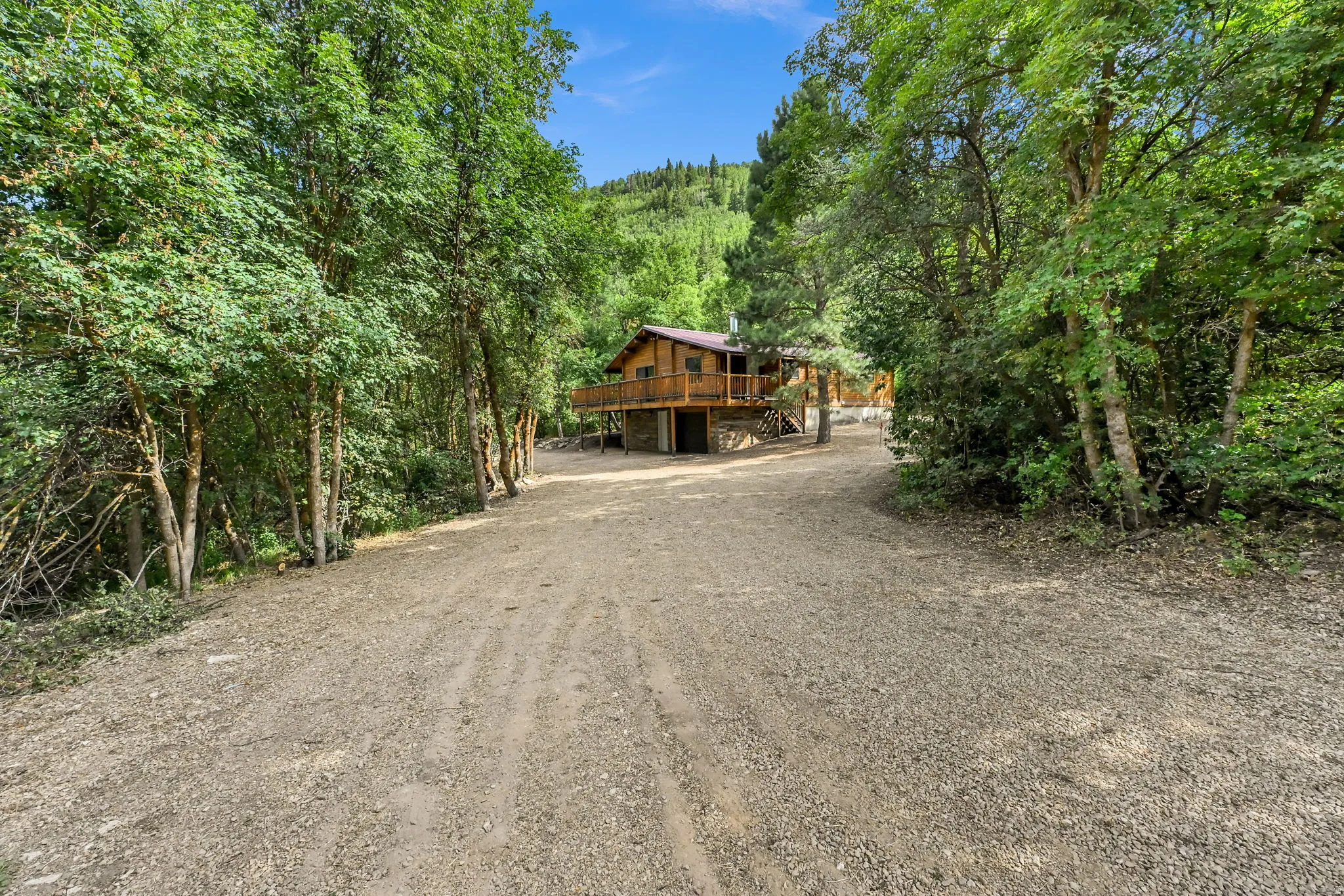 Gravel access road with surrounding mature trees and a rustic wood-sided residence featuring an elevated deck