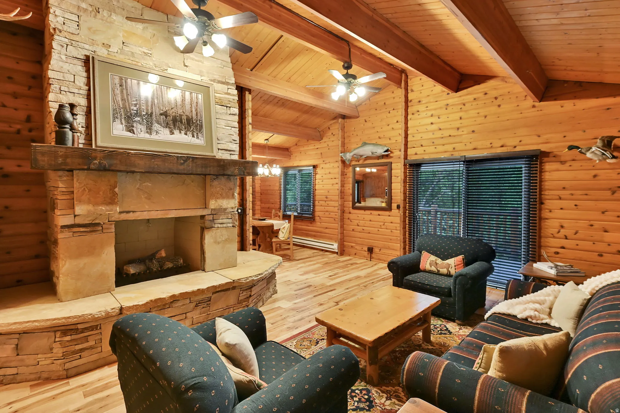 Spacious living area featuring a stone-clad fireplace with a prominent wood mantel, vaulted wood-paneled ceilings with exposed beams, wood-finish flooring, and a large sliding glass door