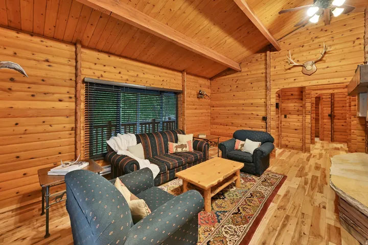 Great room featuring extensive wood plank walls, vaulted wood beam ceilings, wood-finish flooring, a ceiling fan with integrated lighting, and a large window with blinds