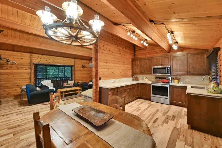 Open-concept living space featuring wood-finish flooring, wood-paneled walls, a vaulted wood-beam ceiling, a wagon wheel chandelier, and a kitchen with wood cabinetry, stainless steel appliances, and light-toned countertops