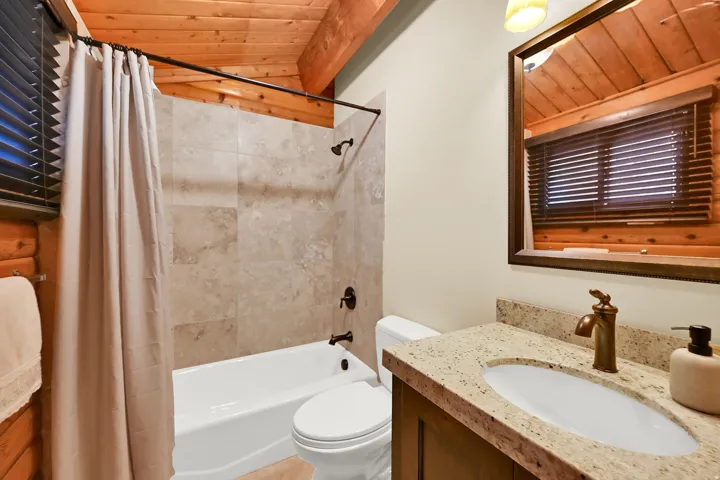 Bathroom featuring a wood-planked ceiling with exposed beams, a tiled shower-over-tub, a vanity with a light-toned countertop, a bronze-finish faucet, and a framed mirror