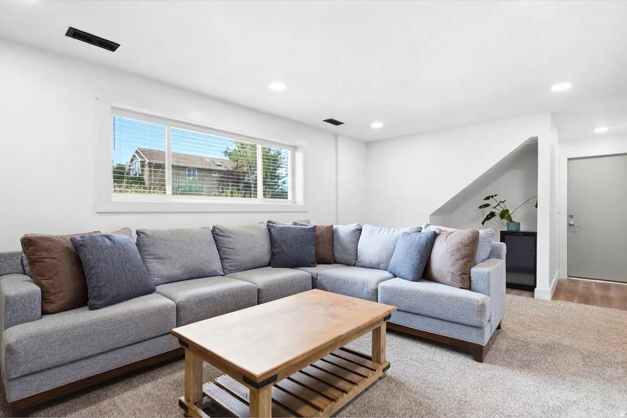 Recessed ceiling lighting, white walls, large window with blinds, light-colored carpeting, and wood-finish flooring