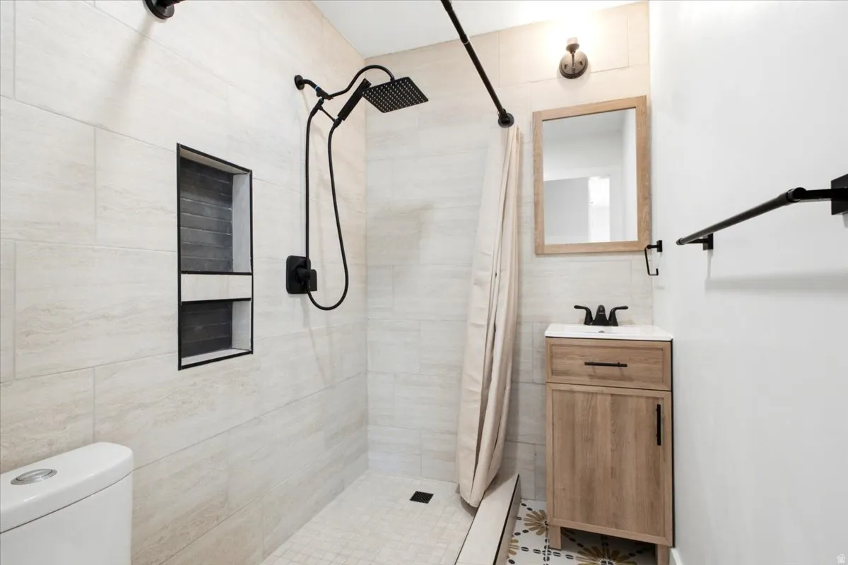 Bathroom featuring a shower with a built-in niche, light-toned wall tiling, black fixtures, wood-finish vanity, and patterned floor tiling