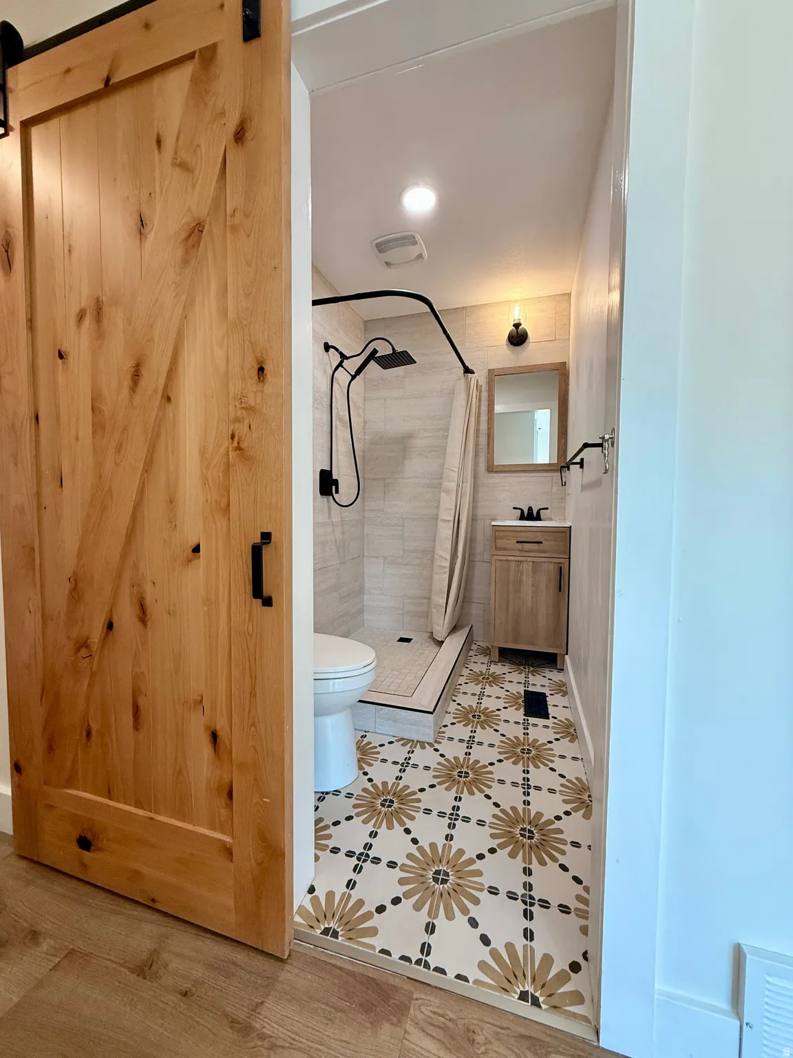 Wood-finish barn door entry to a bathroom featuring patterned floor tiling, a tiled shower with a black fixture, and a wood-finish vanity with a framed mirror