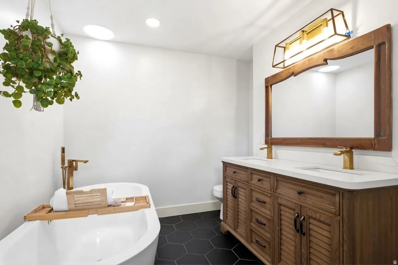 Freestanding soaking tub with a wall-mounted faucet, dual vanity with a light wood finish, white countertop, and dark hexagonal floor tiles