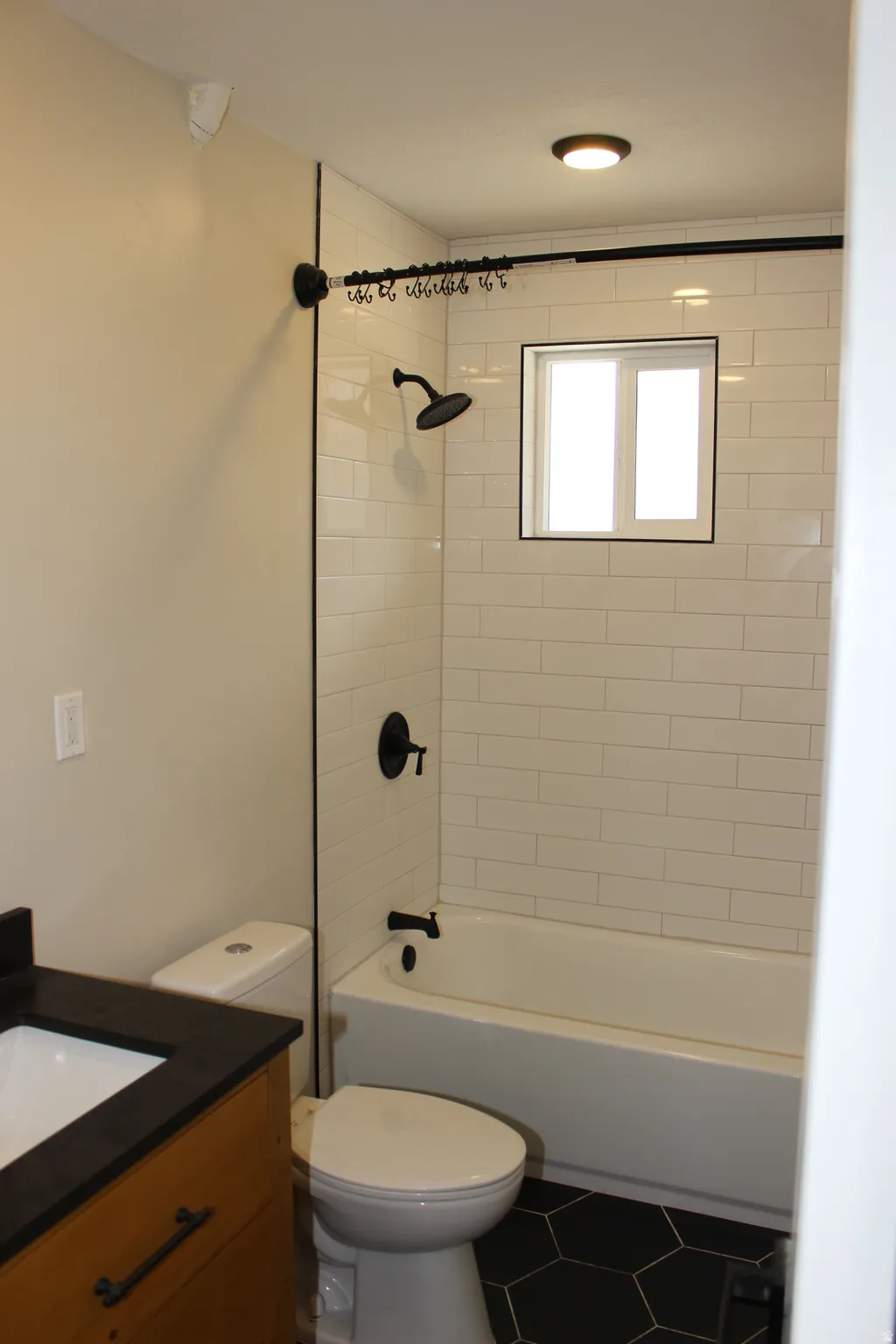 Bathroom featuring a white subway tile shower surround with a built-in window, a white bathtub, a black hexagonal tile floor, a wood-finish vanity with a dark countertop, and matte black fixtures