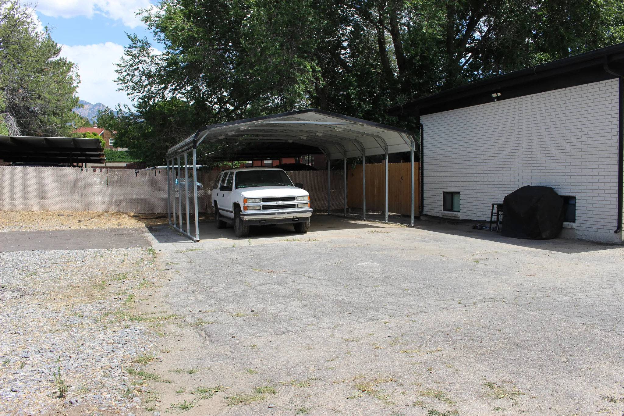 Spacious paved area featuring a metal 2-car carport, a white brick exterior, a black roofline, and mature trees in the background