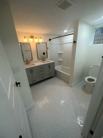 Bathroom featuring herringbone tile flooring, a double vanity with stone countertops, individual framed mirrors, a gold-finish four-light fixture, and a tub/shower combination
