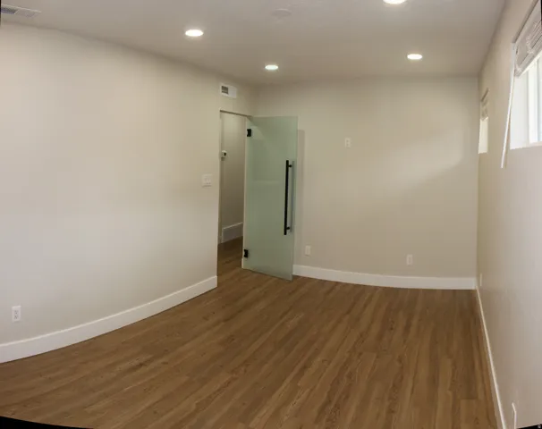 Spacious room featuring wood-finish flooring, recessed ceiling lighting, a frosted glass pivot door with a black pull handle, and multiple windows