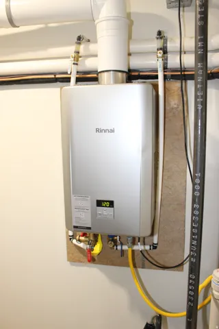 Wall-mounted Rinnai tankless water heater with digital display, visible plumbing, and gas line connections