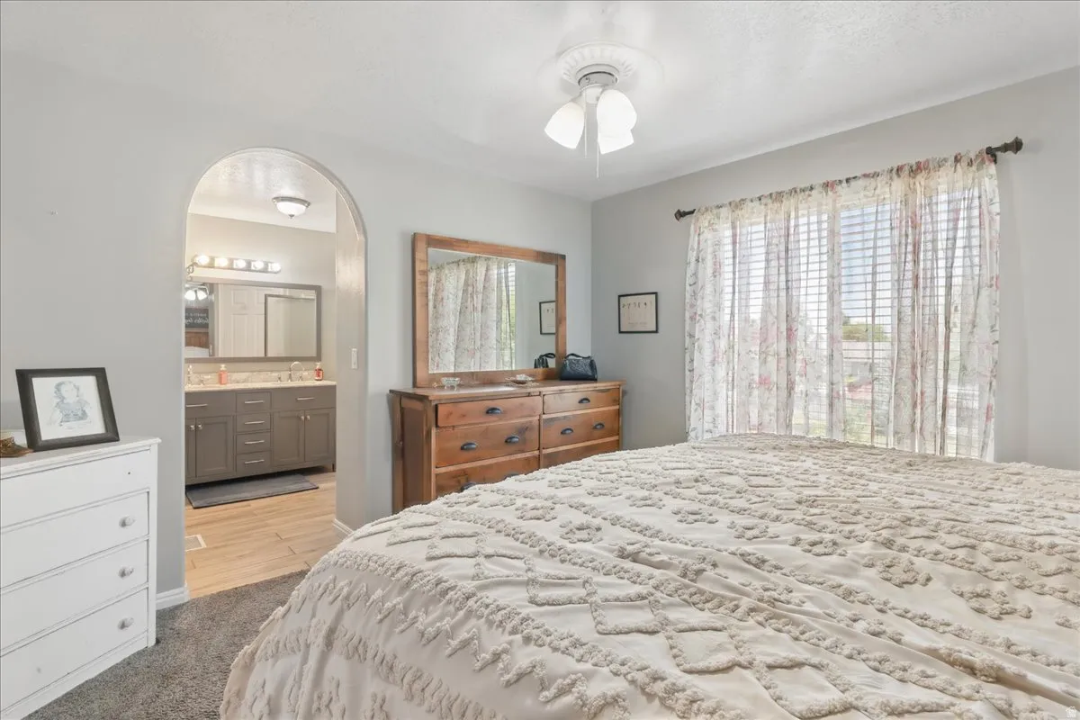 Bedroom featuring a ceiling fan, a large window with horizontal blinds, and an arched doorway leading to a bathroom