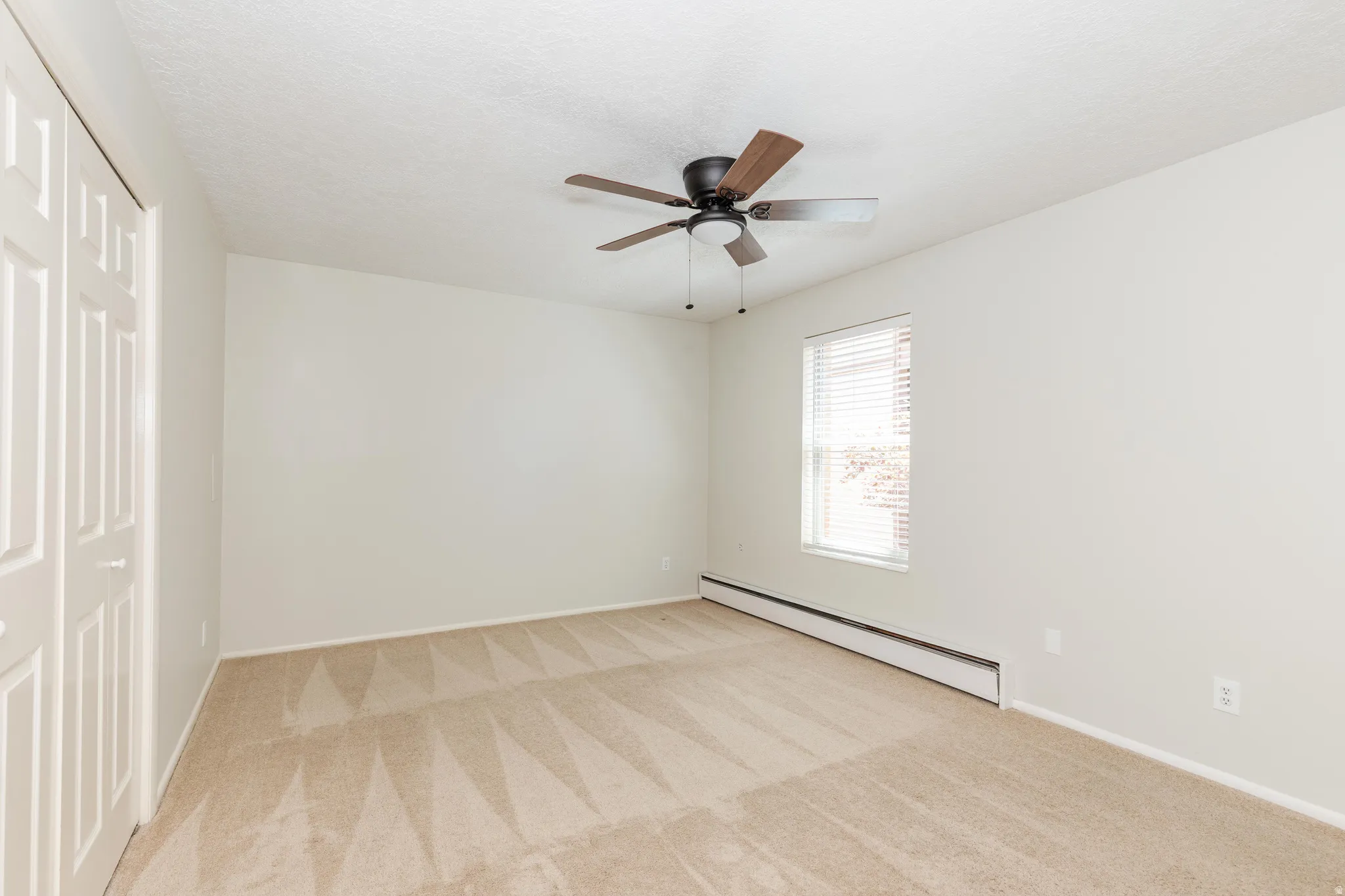 Carpeted room featuring a ceiling fan with wood-finish blades, a single window with blinds, and white bi-fold closet doors