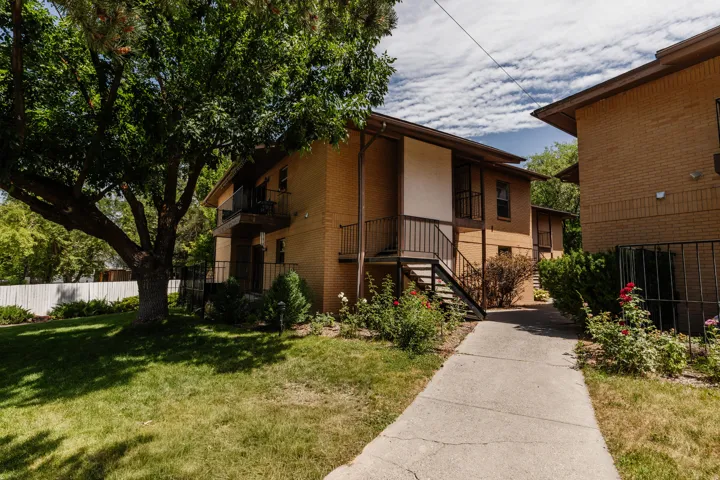 Brick exterior featuring multiple balconies, a concrete walkway, mature landscaping, and a substantial shade tree