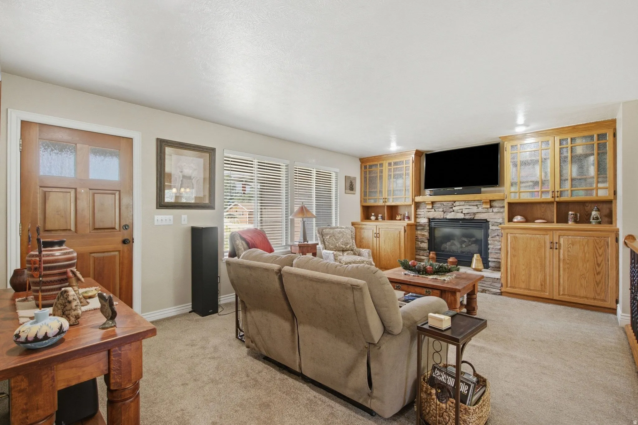 Living area featuring a stone-clad fireplace, integrated wooden cabinetry with glass-front displays, light beige carpeting, multiple windows with blinds, and a solid wood entry door with frosted glass inserts