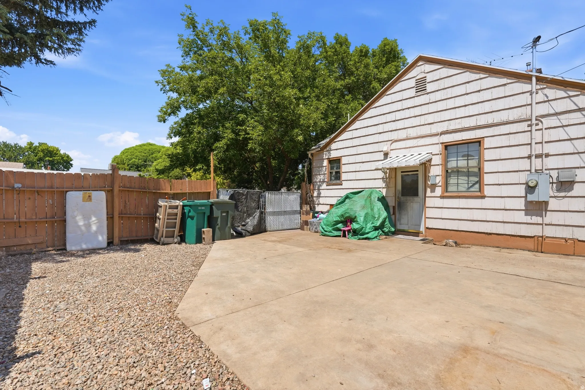 Side exterior of the property featuring white shingle siding, a concrete patio, a gravel area, a wood fence, and mature trees