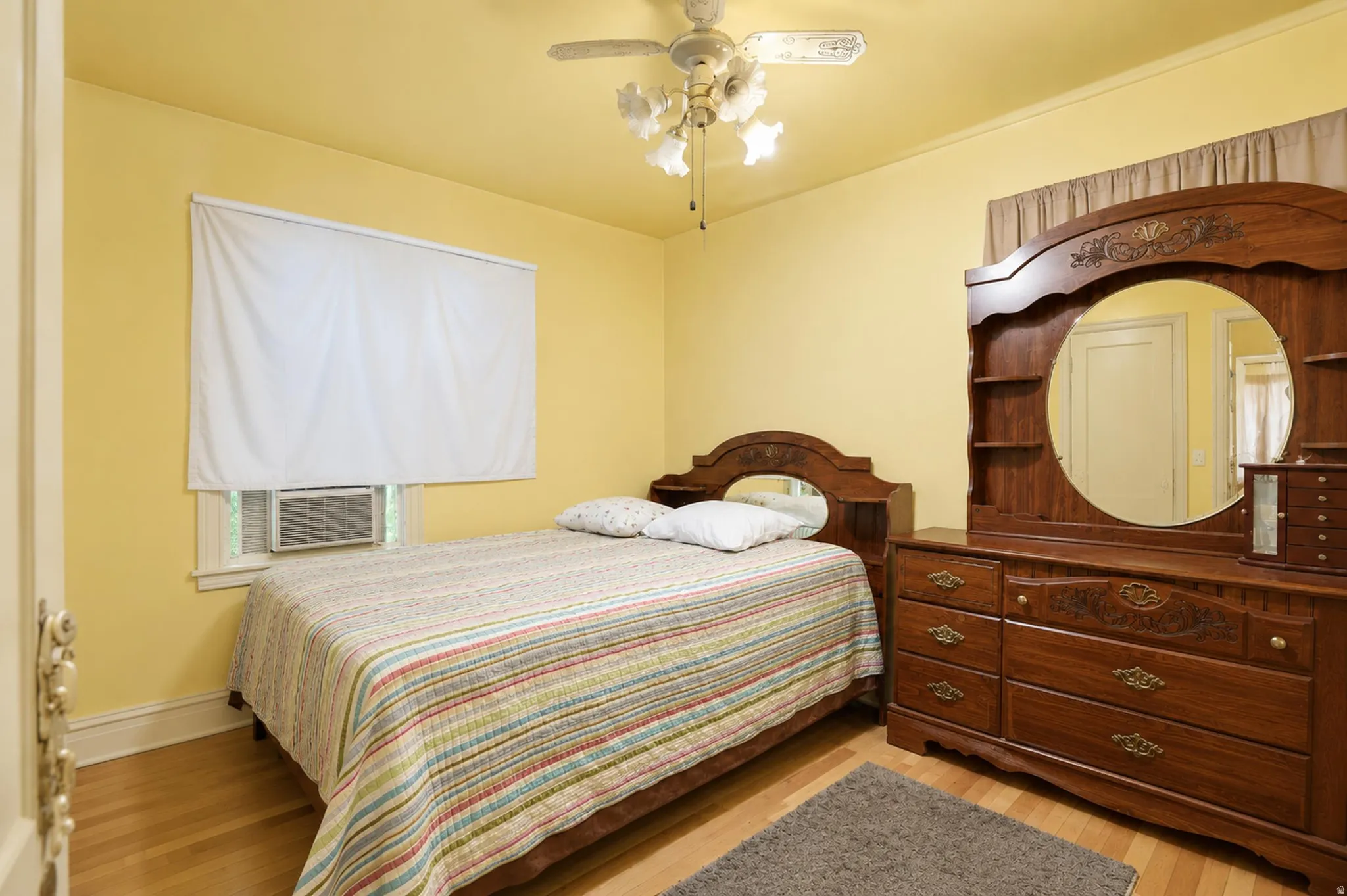 Hardwood flooring throughout, window with white roller shade, ceiling fan with light fixture, window-mounted air conditioning unit
