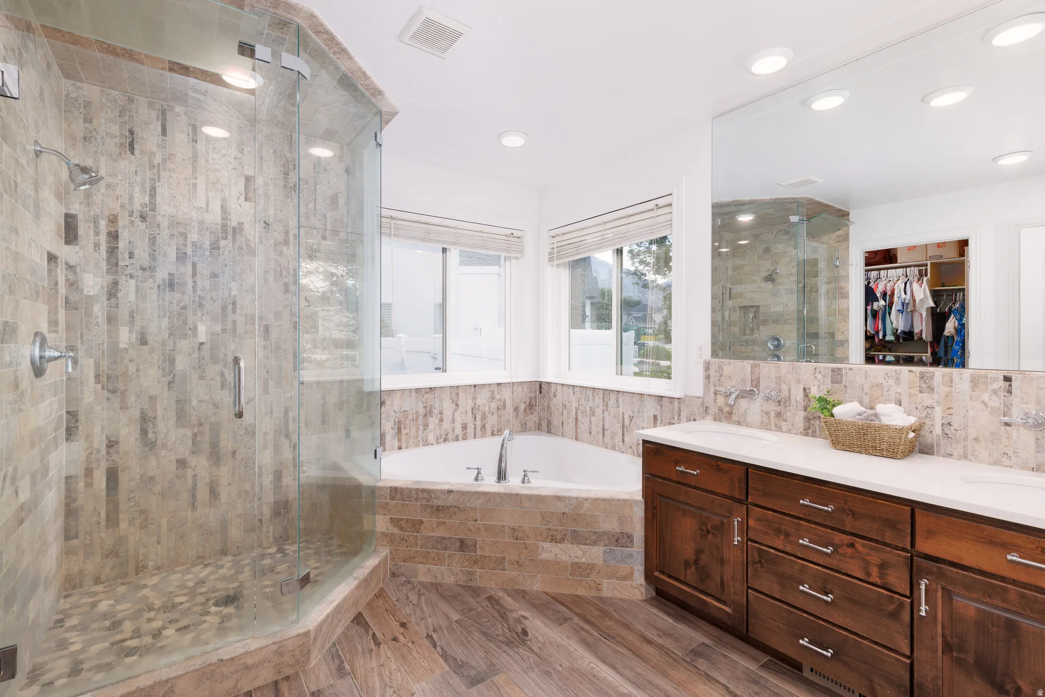 Spacious bathroom featuring a glass-enclosed shower with pebble flooring, a corner soaking tub, dual vanity with white countertops, and wood-finish flooring