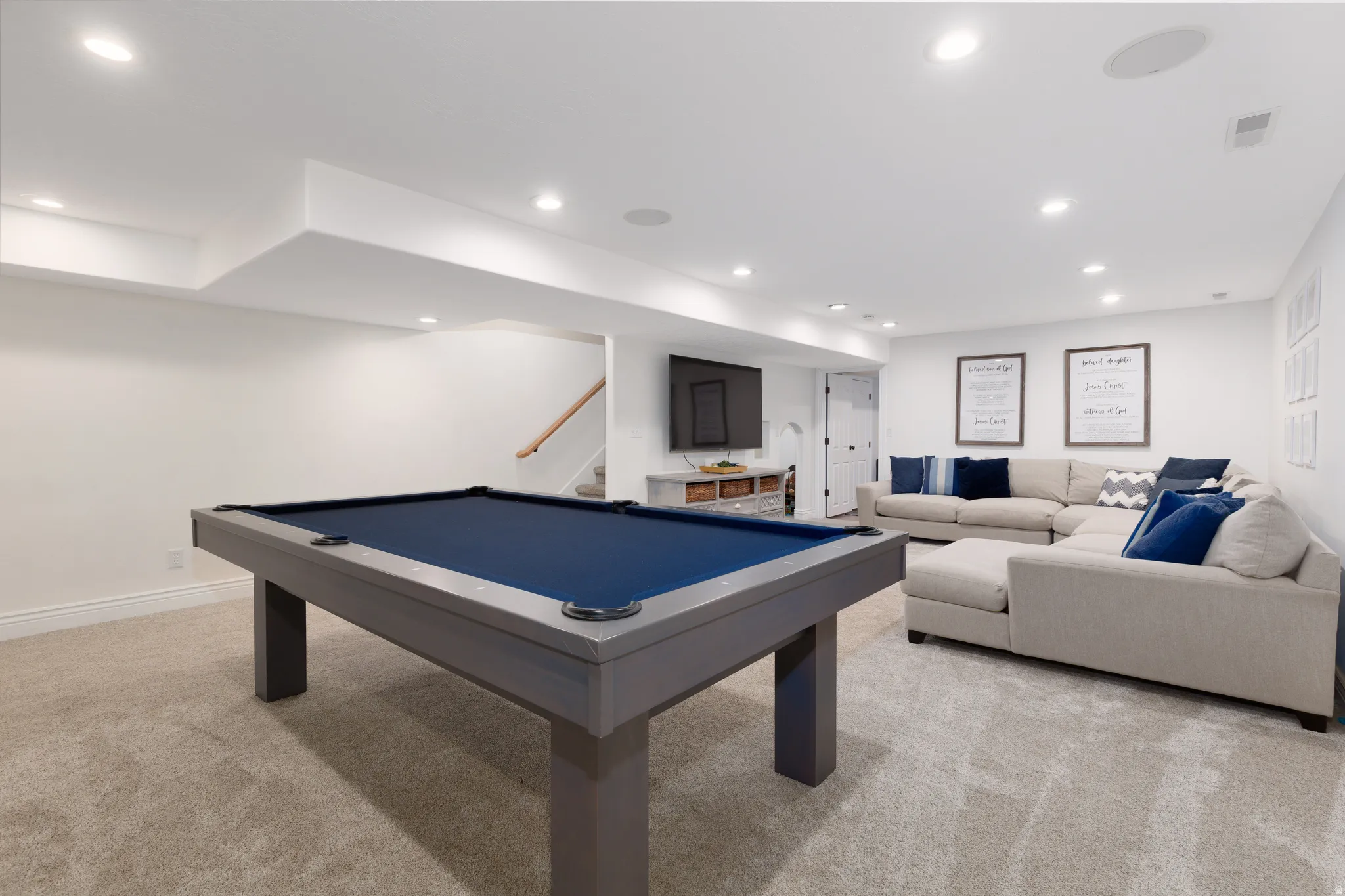 Spacious recreational area featuring recessed lighting, light-toned carpeting, white walls, a visible staircase with wood railing, and a sectioned sofa