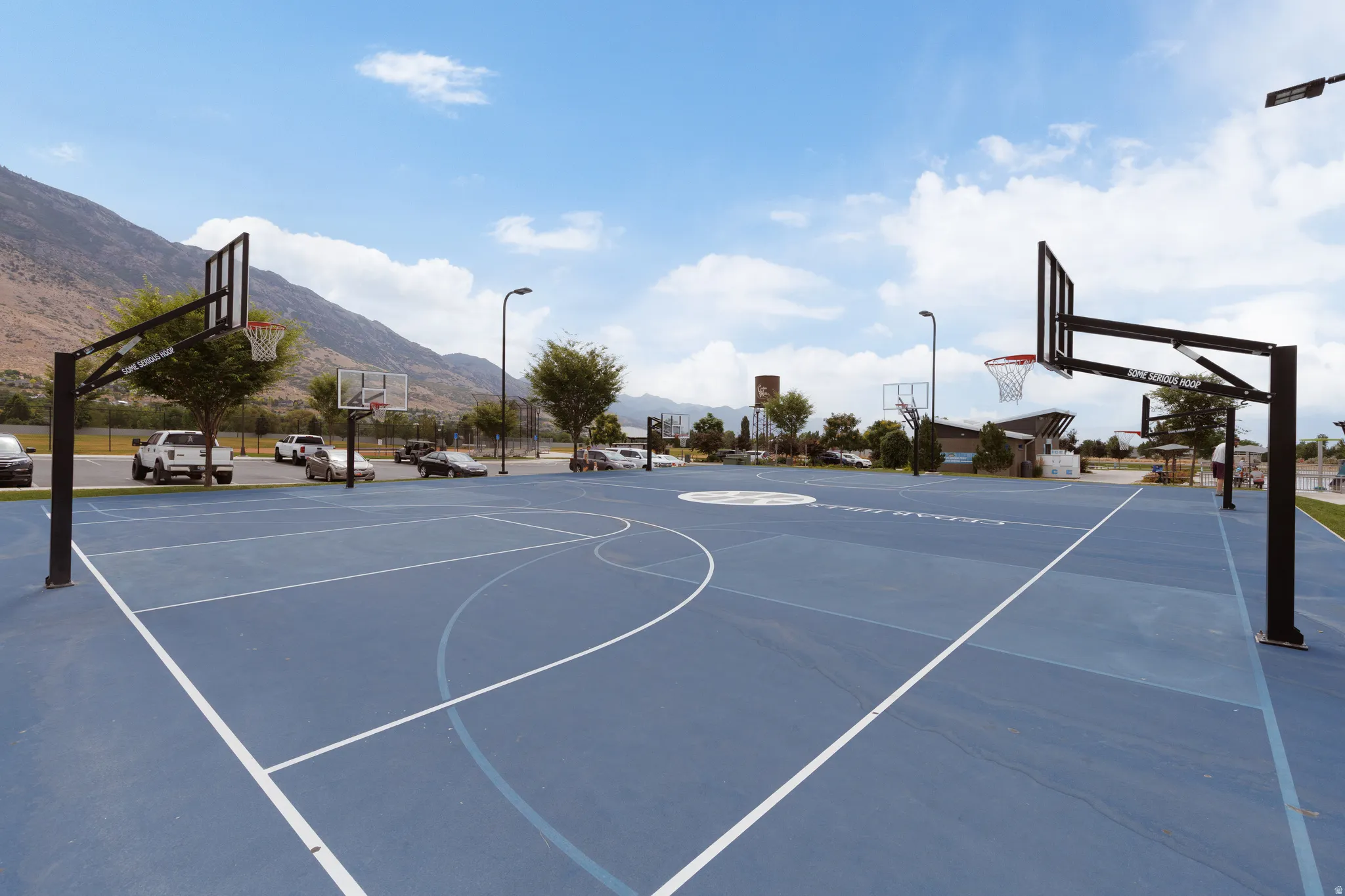 Full-sized basketball court featuring a blue surface with white markings, multiple hoops with black support structures, and perimeter lighting