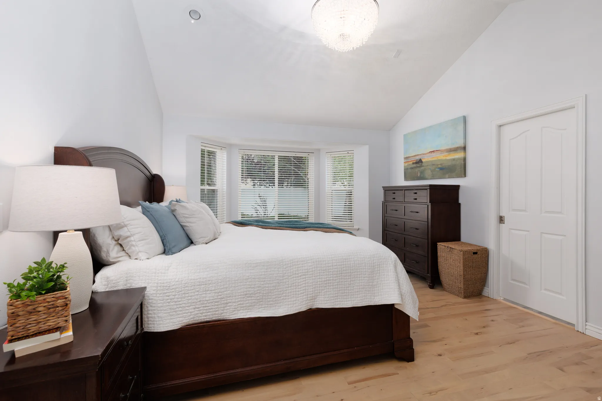 Vaulted ceiling bedroom featuring light wood-finish flooring, a bay window with blinds, a white paneled door, and a decorative ceiling light fixture