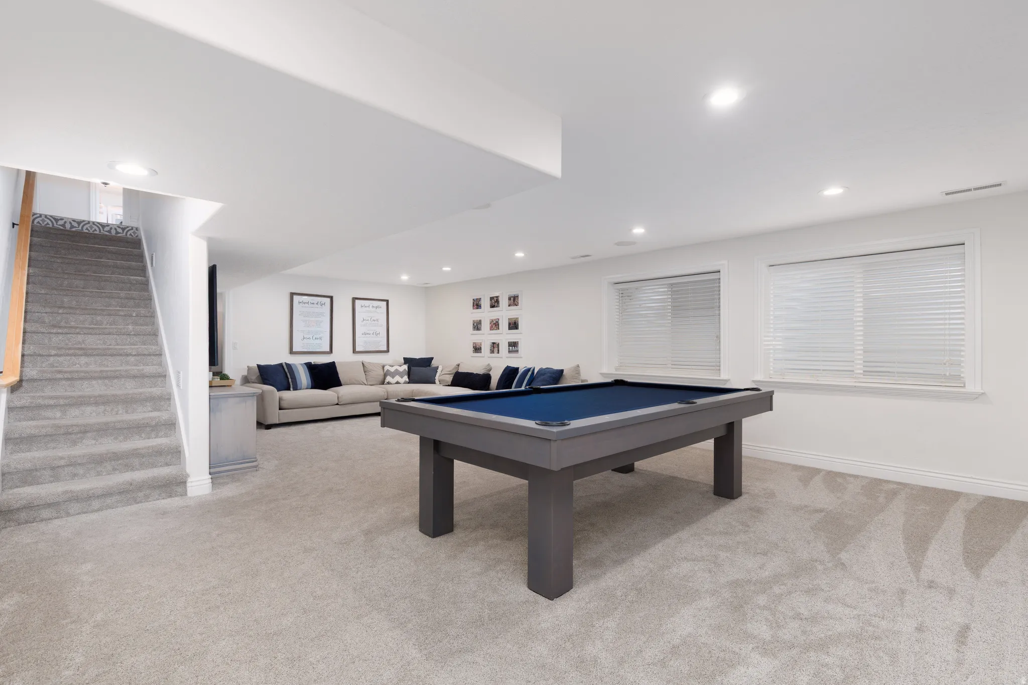 Spacious carpeted room featuring a built-in staircase with wood handrail, recessed lighting, and multiple windows with blinds