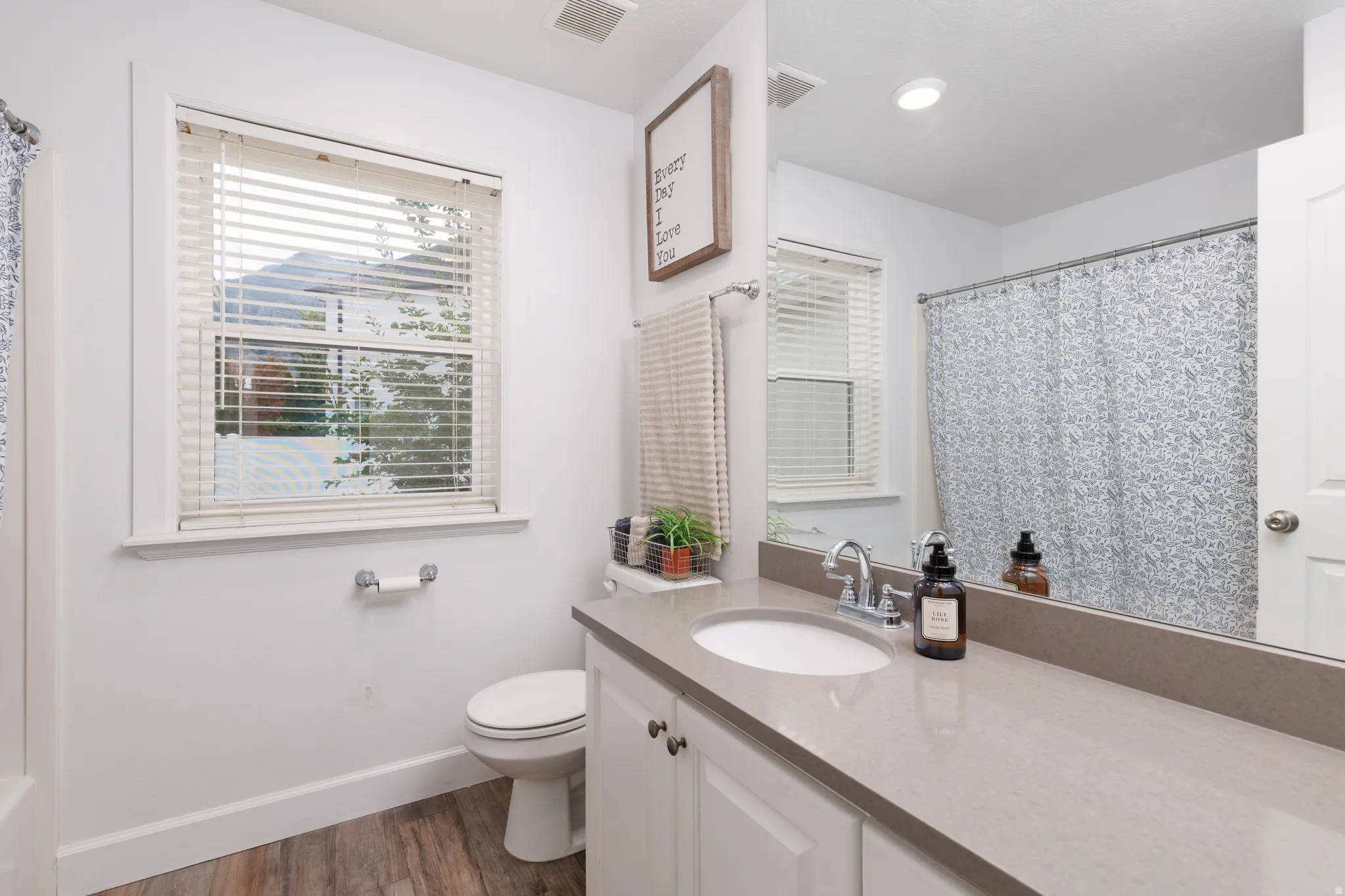 Bathroom featuring a window with blinds, wood-finish flooring, a white vanity with a light-colored countertop, and a built-in shower/tub with a patterned curtain