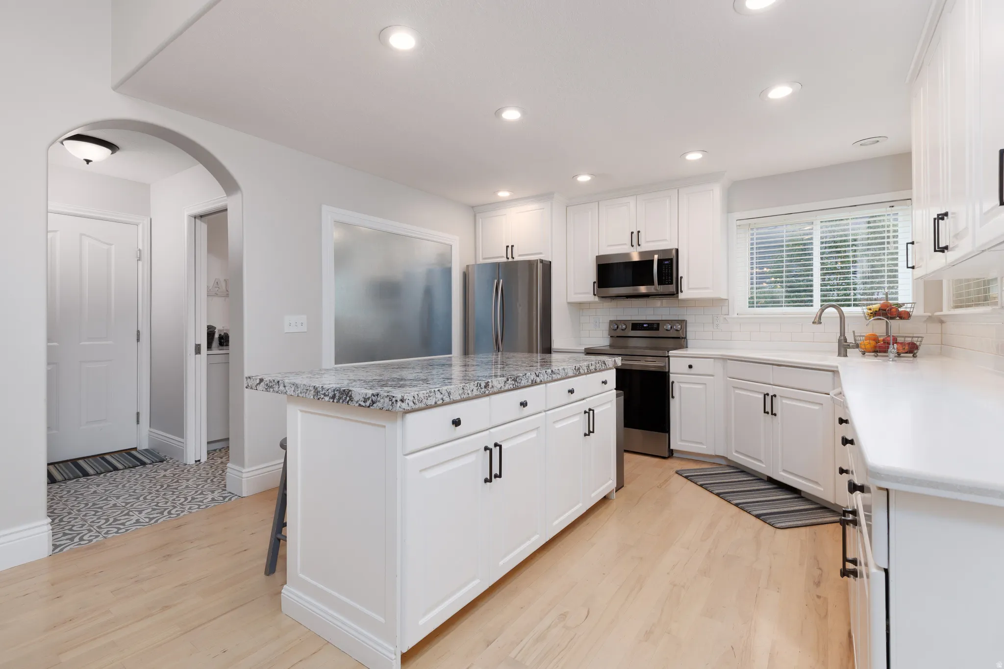 Contemporary kitchen featuring white cabinetry, light wood-finish flooring, a central island with a stone-finish countertop, stainless steel appliances, and a tiled backsplash